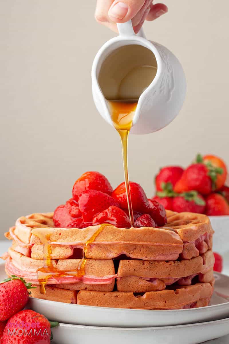 homemade strawberry waffles stacked on a plate with maple syrup being poured on top.