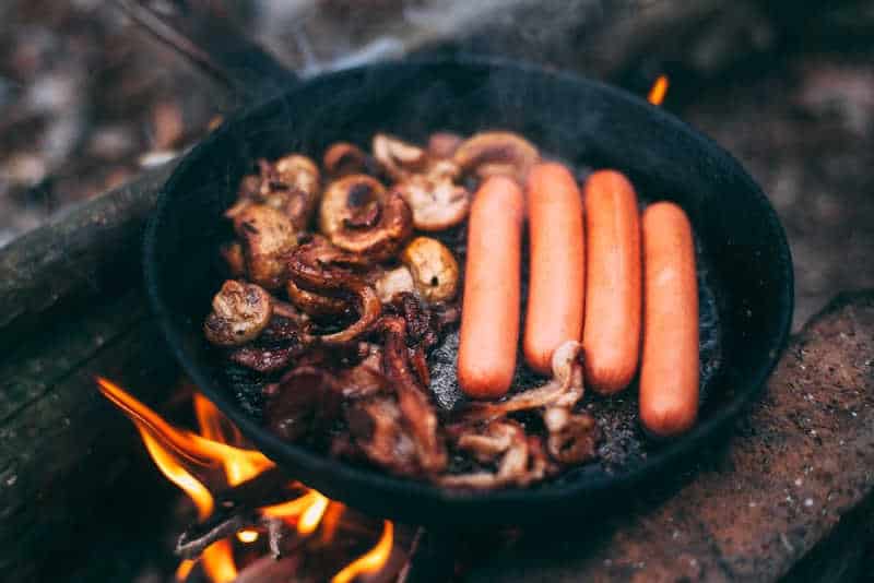 Fried mushrooms and bacon with sausages in a pan in the woods on fire. Food in nature. Cooking food on the road. Picnic