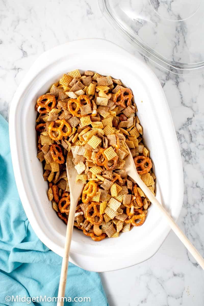 white crockpot filled with the ingredients to make slow cooker chex mix with 2 spons mixing it