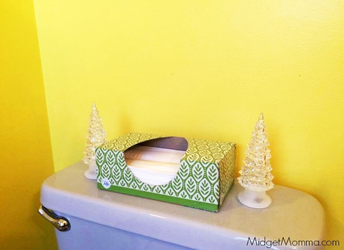 Bathroom themed Kleenex box sitting between 2 white plastic Christmas tree decorations on the back of a toilet.