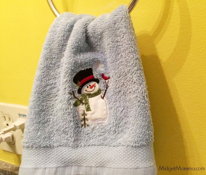 Light blue hand towel with a snowman on it, hanging from a towel ring in a bathroom, on a yellow wall.