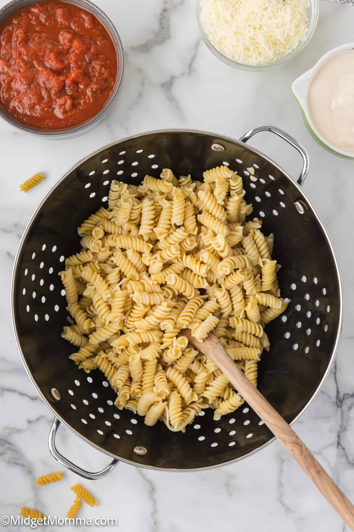 Cooked pasta in a colander with a wooden spoon, with grated cheese, tomato sauce, and cream in separate containers nearby.