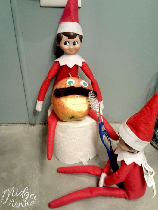 Elf on the shelf with a tooth brush, brushing an apple character with a funny mustache