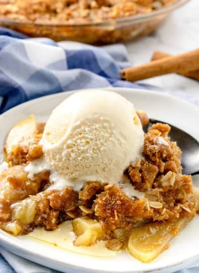 Apple crisp topped with ice cream on a plate with cinnamon sticks and apples