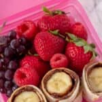 Nutella And Banana Sushi roll up in a pink lunch box container with grapes, strawberries and raspberries