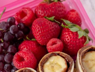 Nutella And Banana Sushi roll up in a pink lunch box container with grapes, strawberries and raspberries
