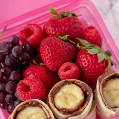 Nutella And Banana Sushi roll up in a pink lunch box container with grapes, strawberries and raspberries