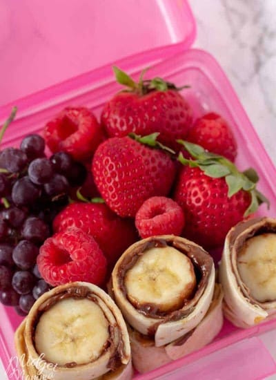 Nutella And Banana Sushi roll up in a pink lunch box container with grapes, strawberries and raspberries