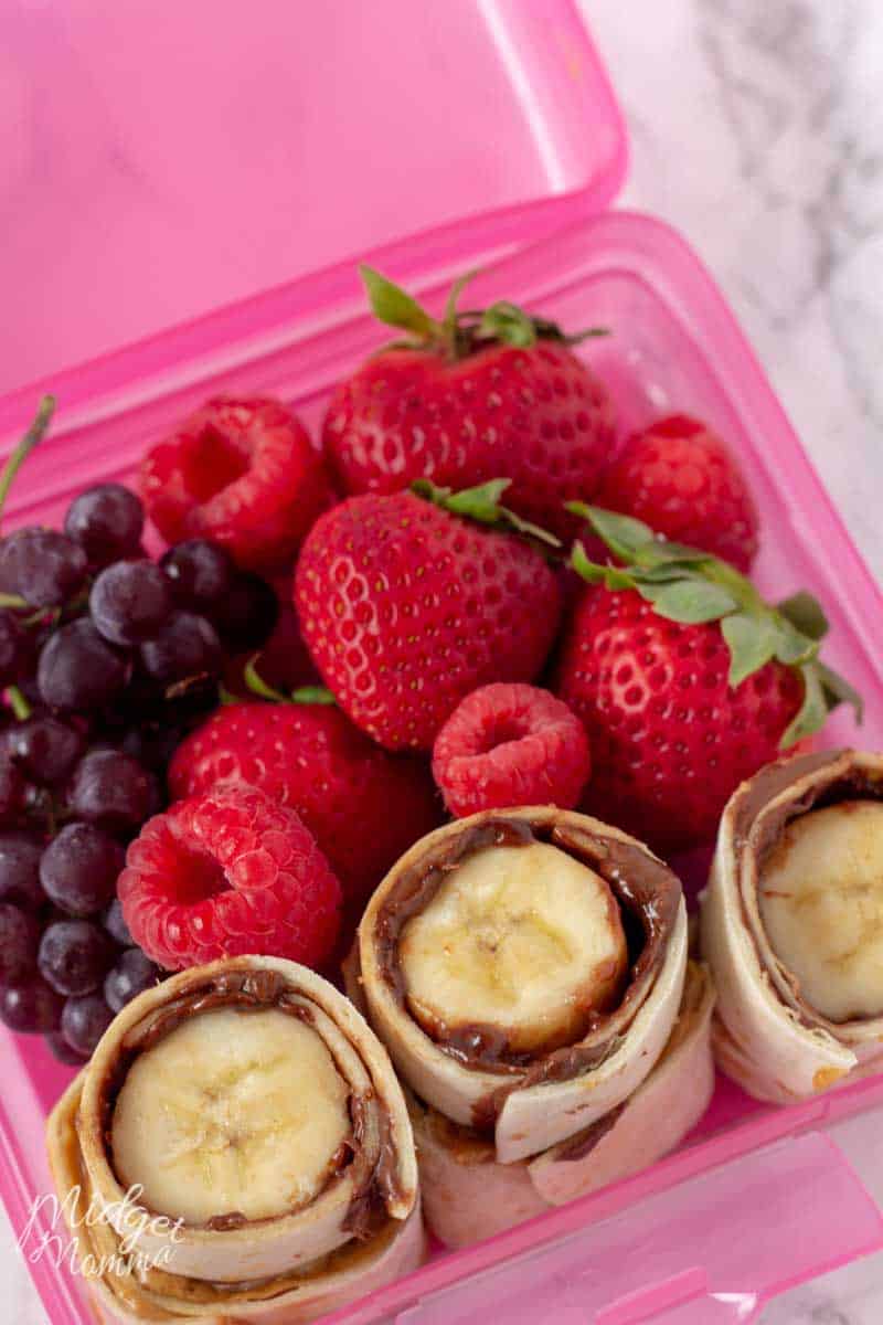 Nutella And Banana Sushi roll up in a pink lunch box container with grapes, strawberries and raspberries