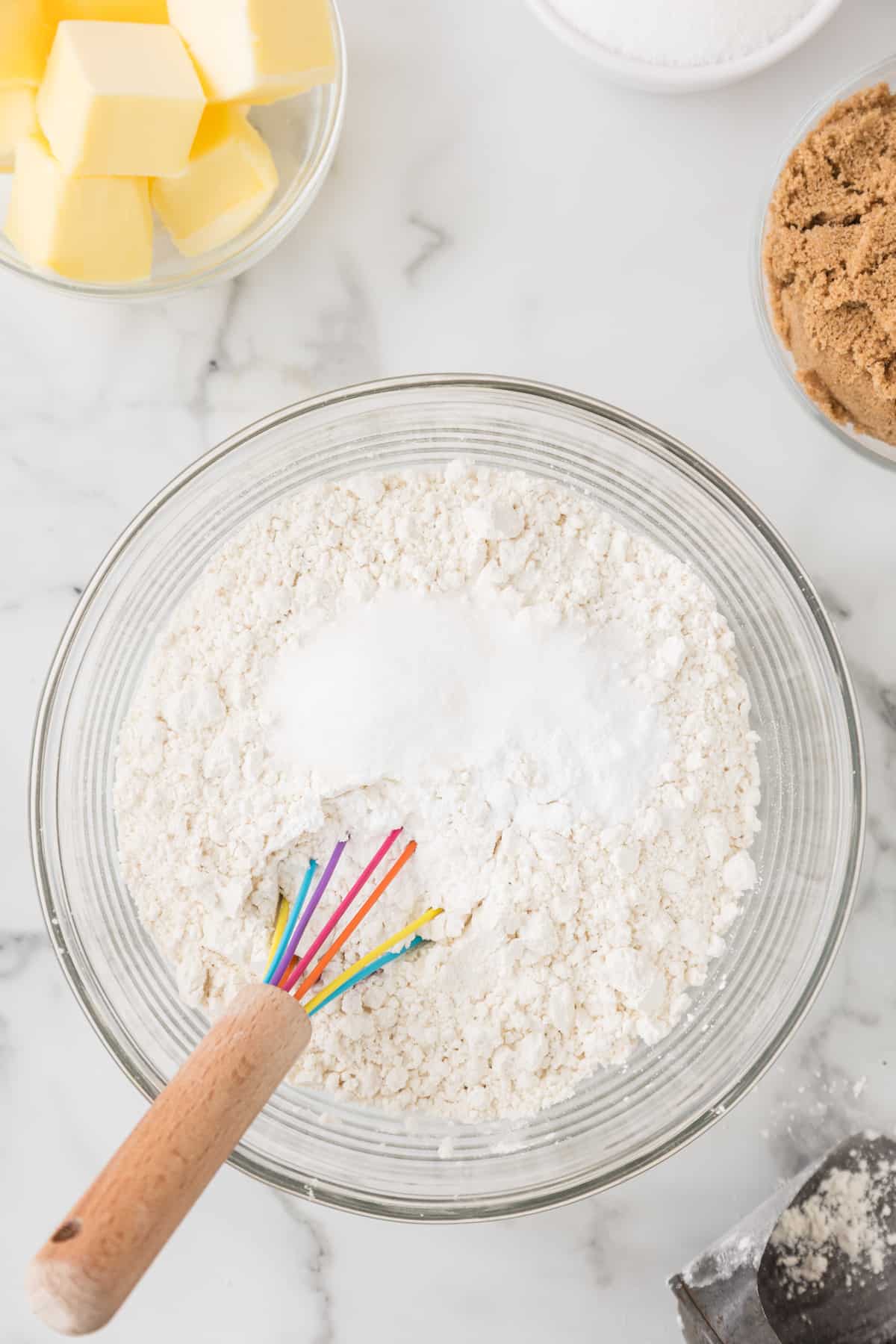 A bowl of flour, eggs, sugar, and butter next to a whisk.