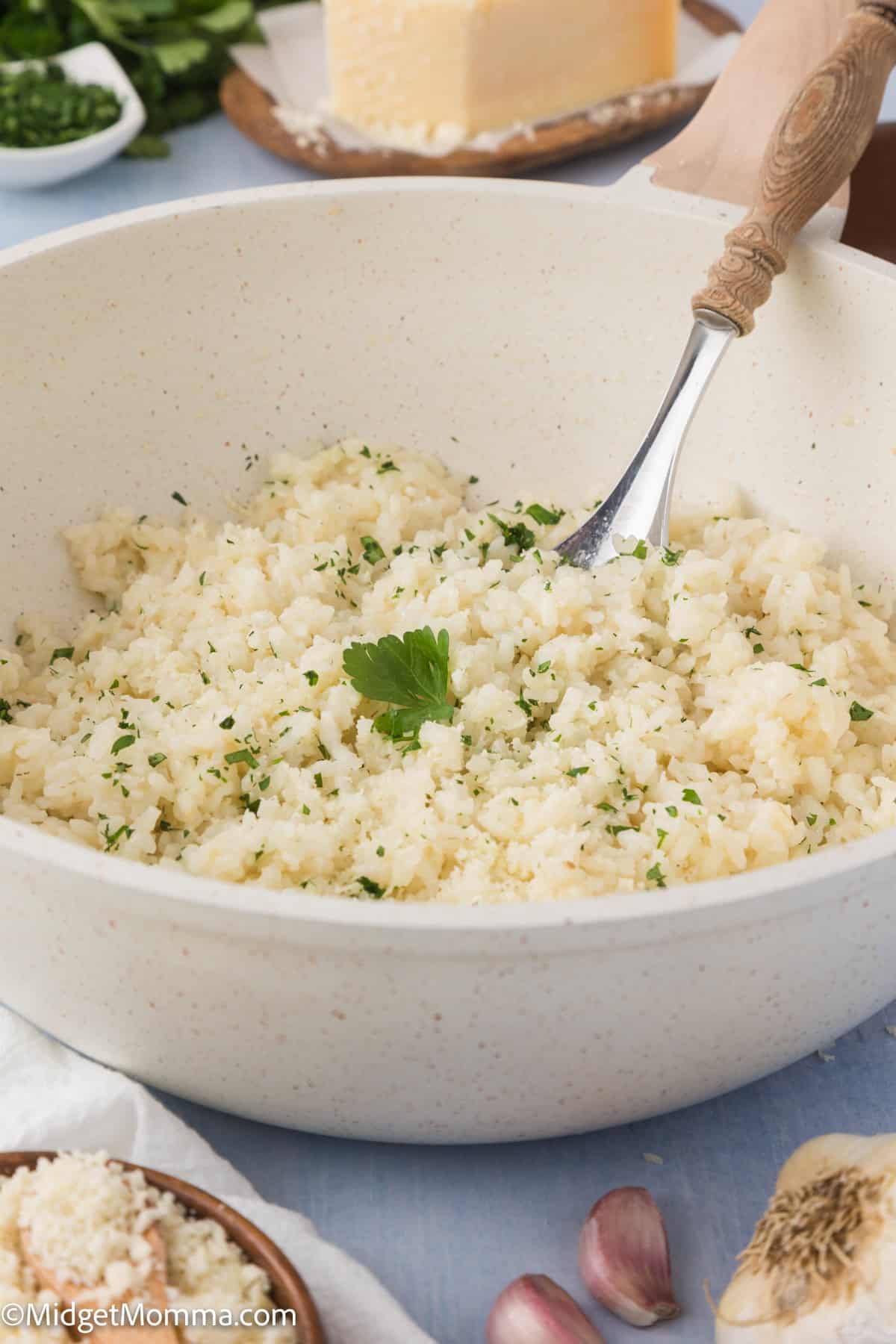 A bowl of creamy parmesan rice garnished with chopped parsley, with a fork in it, surrounded by ingredients like garlic and cheese on a blue tablecloth.