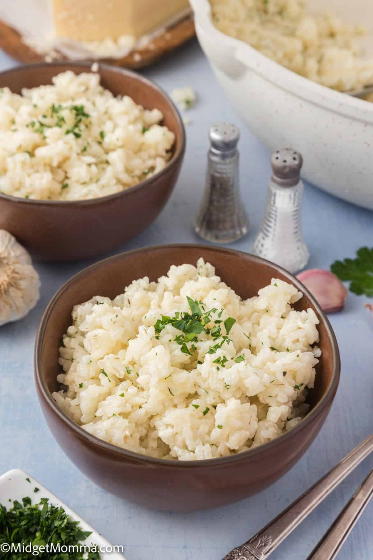 Two bowls of creamy rice garnished with herbs on a blue tablecloth, with garlic cloves and a cheese wheel in the background.