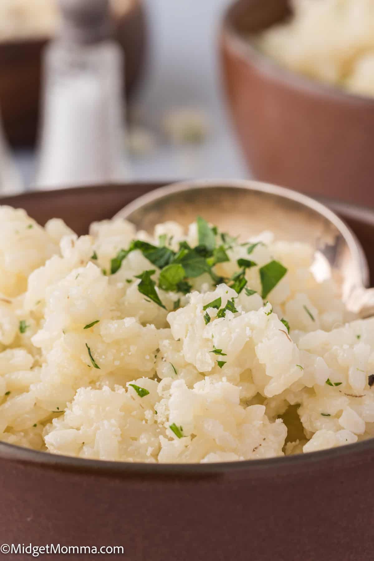A bowl of parmesan rice garnished with chopped parsley, with a silver spoon, on a table with salt shaker visible in the background.