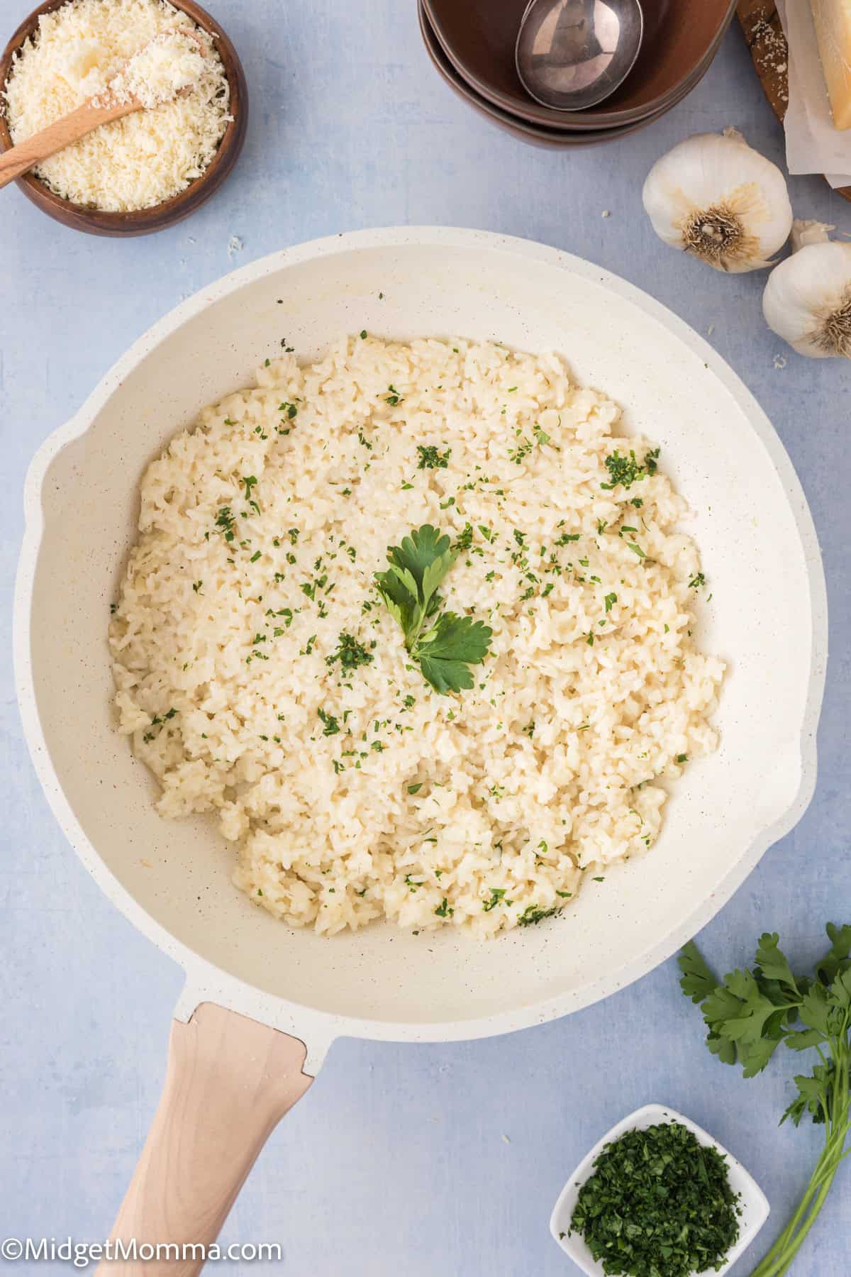 A bowl of creamy rice garnished with parsley, surrounded by ingredients like garlic,  herbs on a light blue surface.