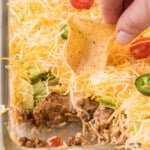 A person dipping a tortilla chip into a casserole dish of layered taco dip