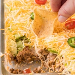 A person dipping a tortilla chip into a casserole dish of layered taco dip