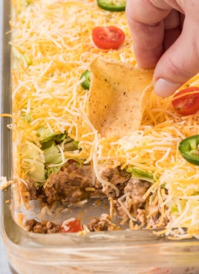 A person dipping a tortilla chip into a casserole dish of layered taco dip