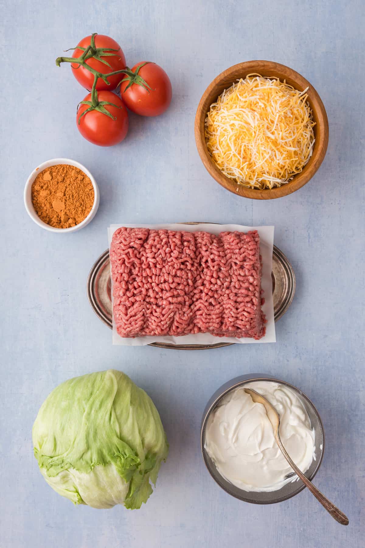 The ingredients for a layered taco dip-  cheese, tomatoes, lettuce and sour cream on a blue background.