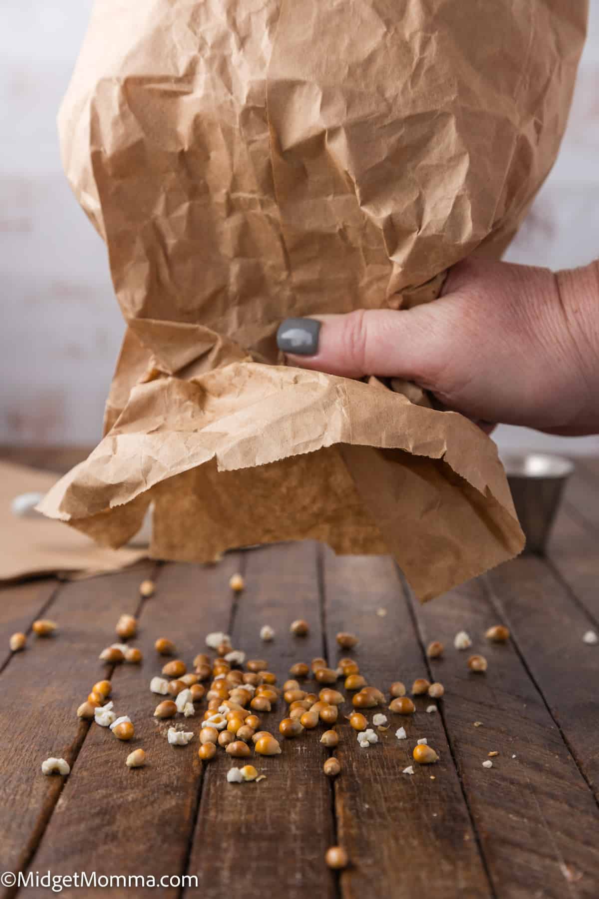 A hand tipping a paper bag and spilling popcorn kernels onto a wooden surface.