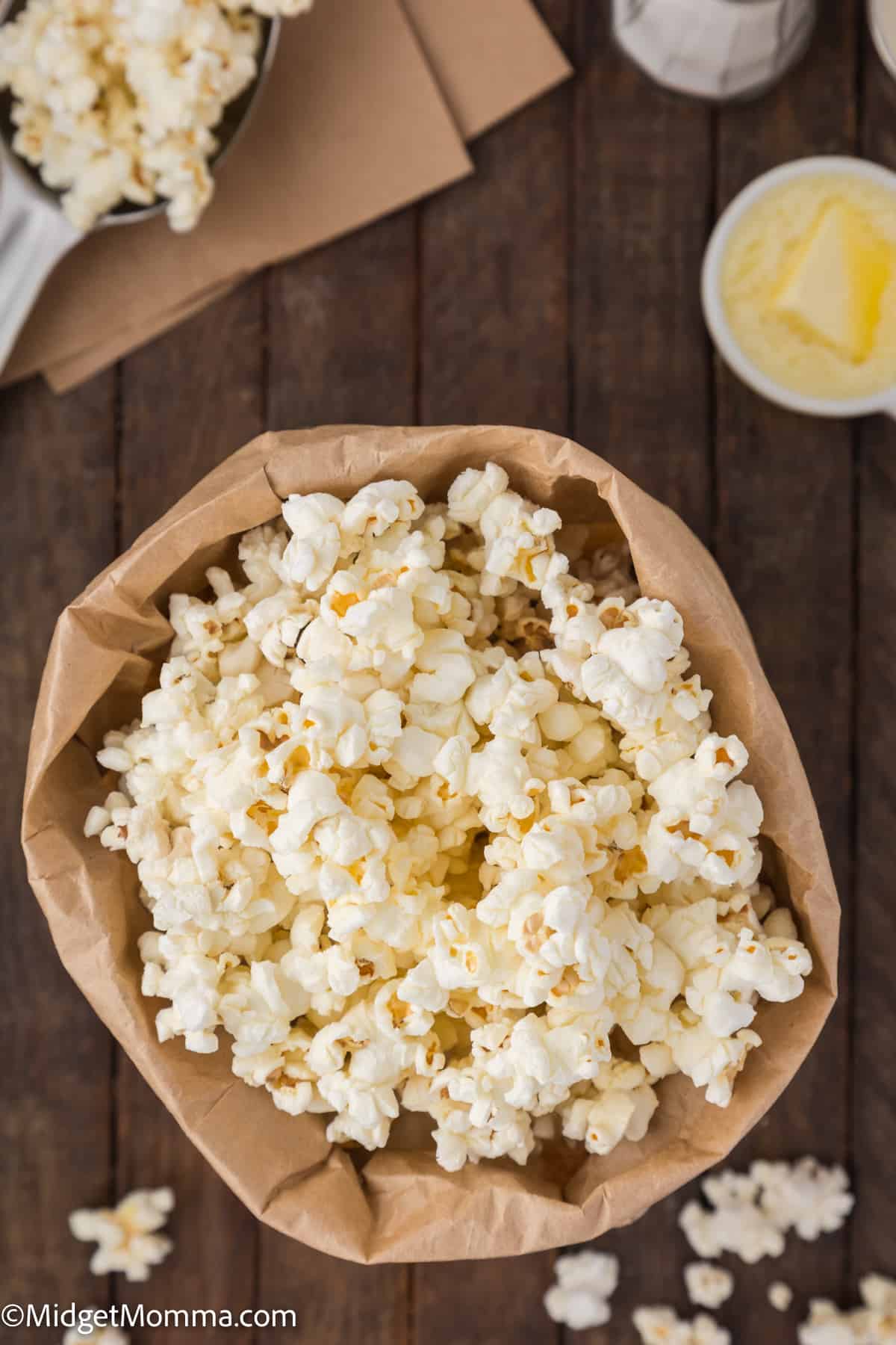 A brown paper bag filled with popcorn on a wooden surface, with more popcorn in an open cardboard box and a bowl of melted butter nearby.