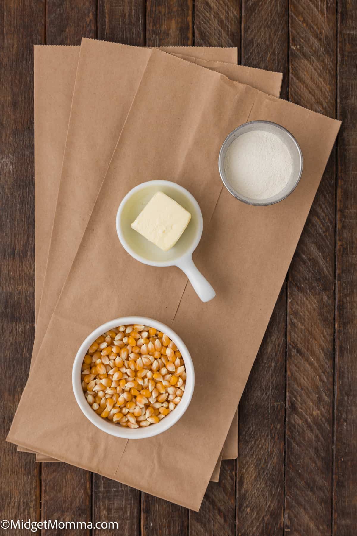Ingredients for making brown bag microwave popcorn arranged on a wooden surface: a bowl of popcorn kernels, a bowl of salt, a stick of butter.