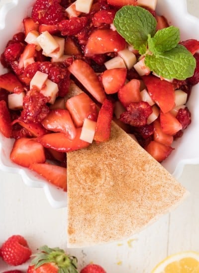 a bowl of fruit salsa with strawberries and raspberries with a cinnamon tortilla chip being dipped in