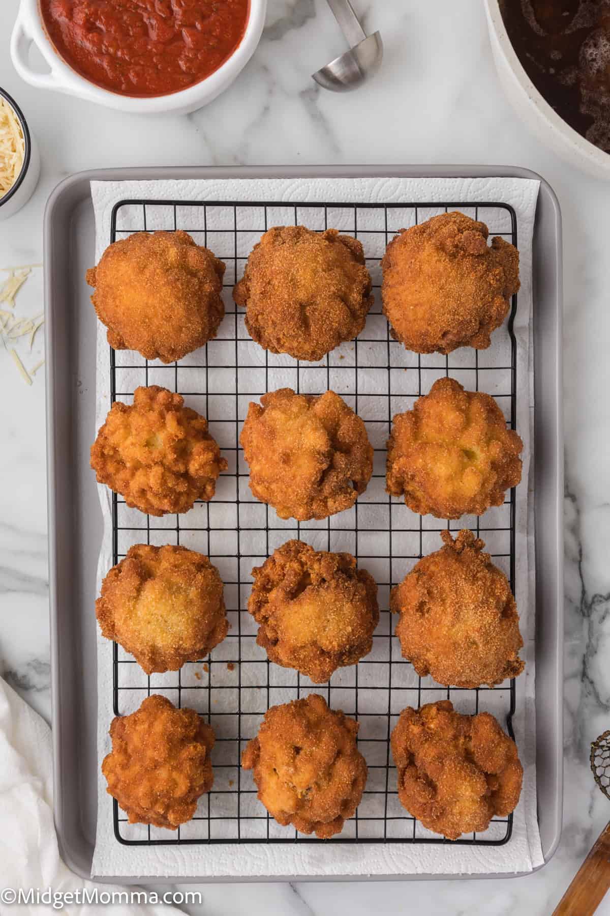 Freshly fried mac and cheese balls arranged on a cooling rack over a baking sheet, with marinara sauce and a spoon visible in the background.