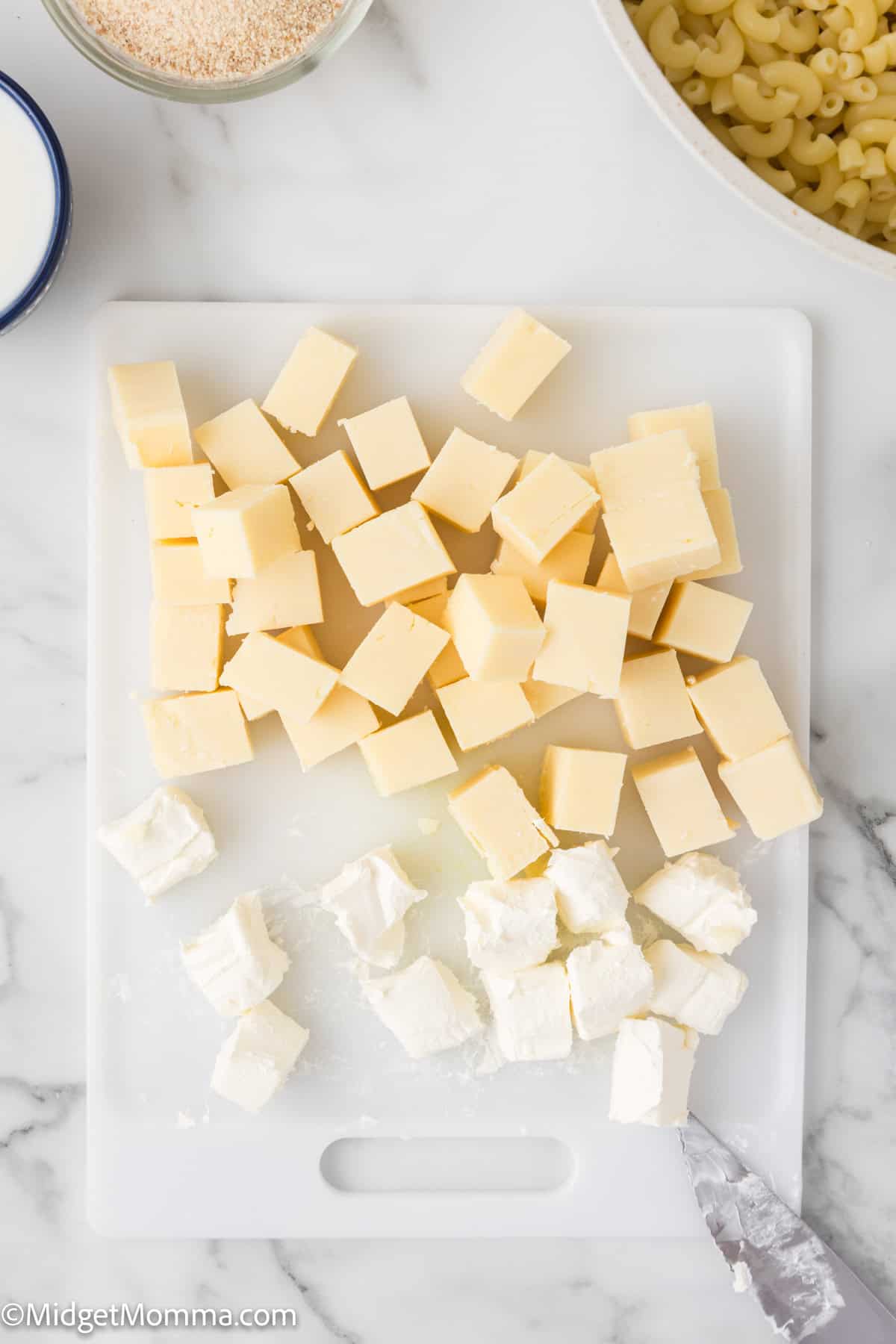 Chopped cubes of yellow cheddar and cream cheese on a white cutting board, with a knife and ingredients like pasta and flour in the background.