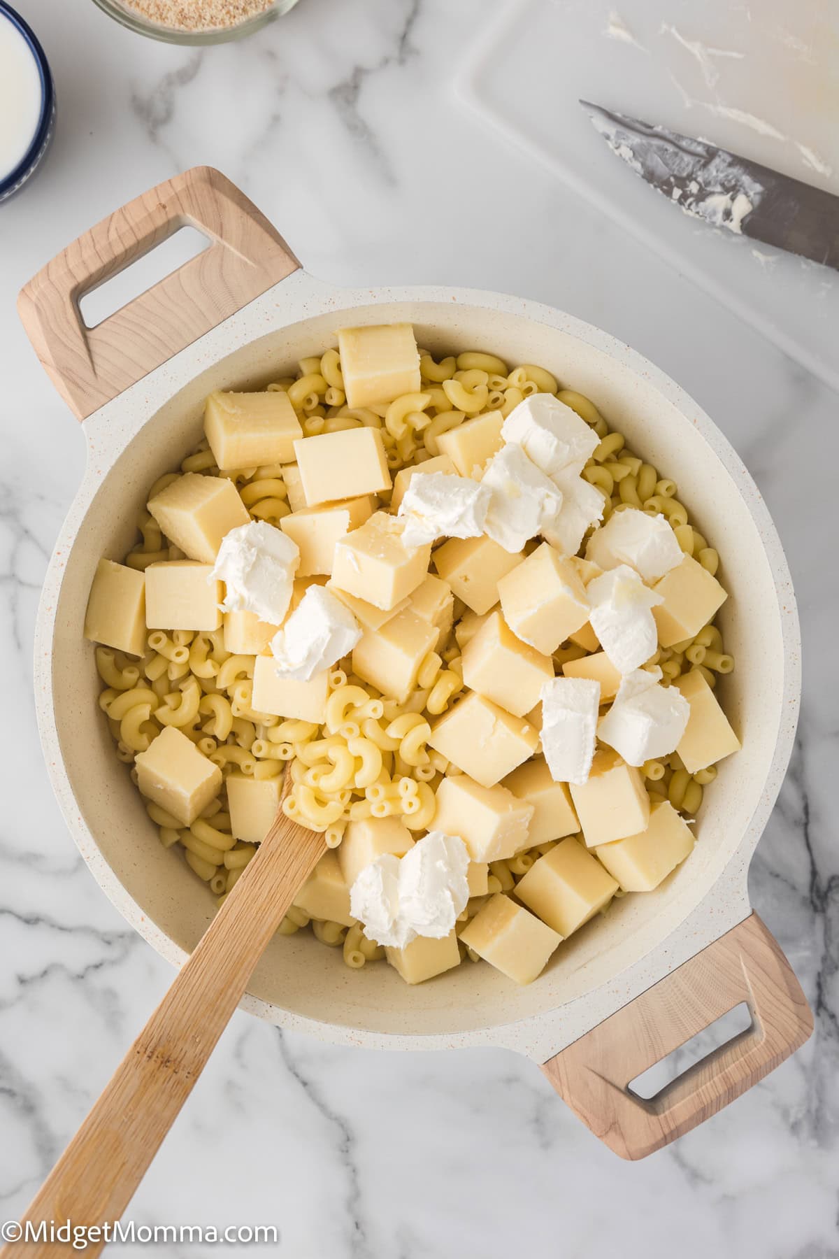 A pot containing cooked macaroni mixed with various types of cheese, with a wooden spoon, on a marble countertop.