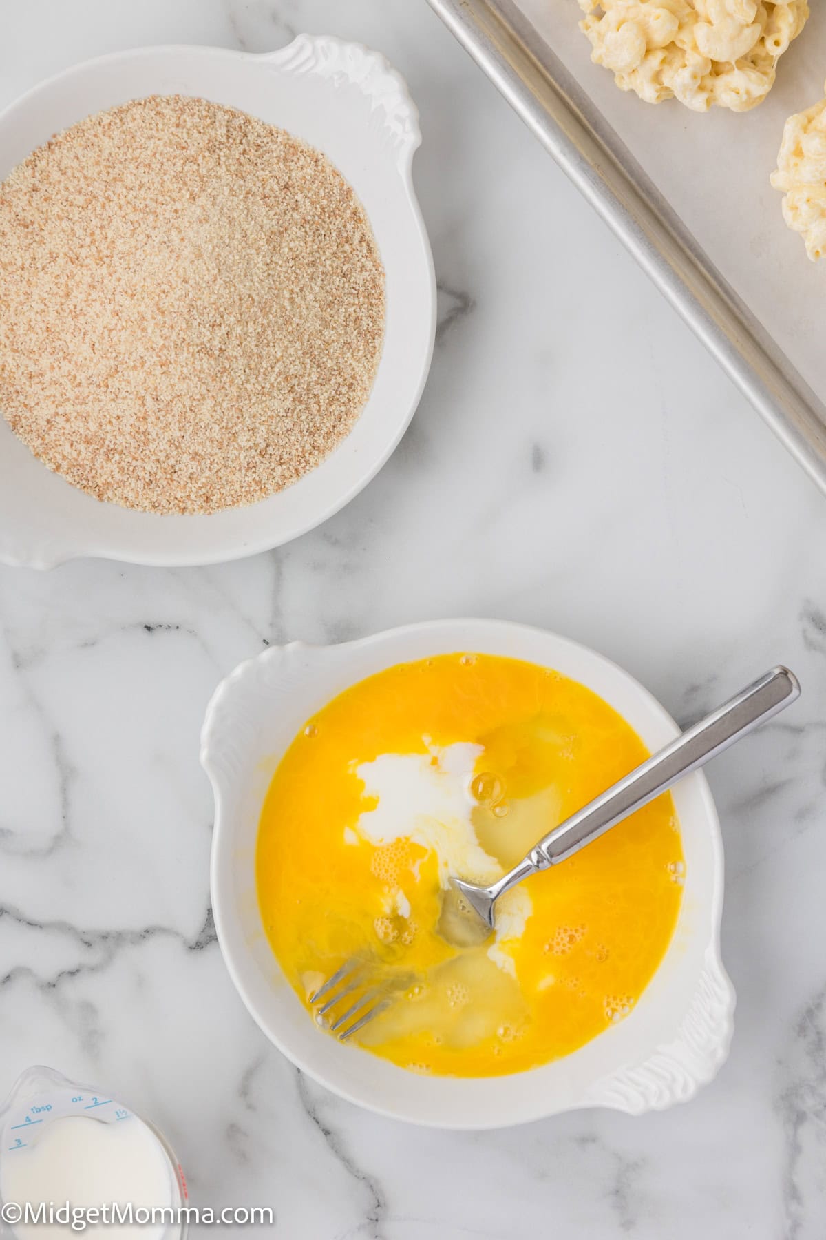 A bowl of beaten eggs alongside a bowl of breadcrumbs, prepared for cooking, on a marble surface.