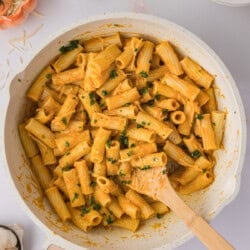 A bowl of creamy pumpkin sauce pasta topped with fresh herbs, surrounded by garlic cloves with a wooden spoon.