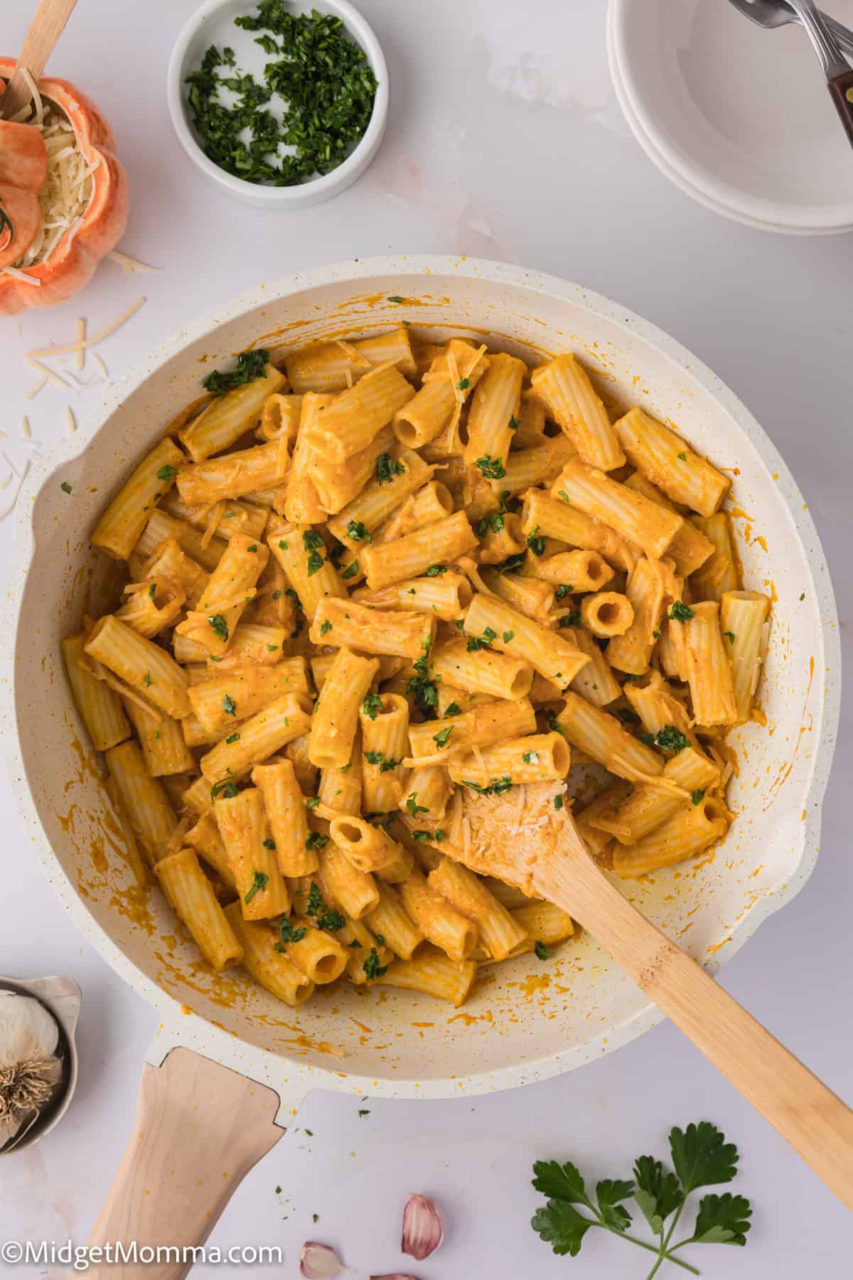 A bowl of pasta topped with a creamy pasta sauce and topped with fresh herbs, surrounded by garlic cloves with a wooden spoon.