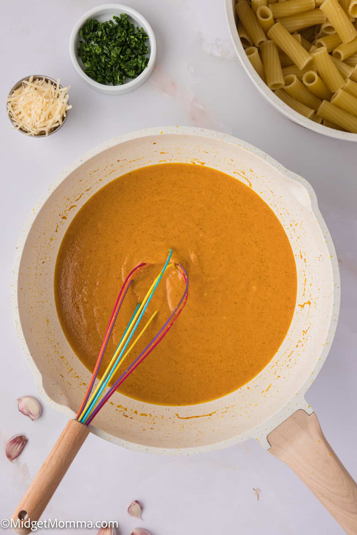 A pot of creamy pumpkin sauce with a colorful whisk, surrounded by ingredients including pasta, garlic, cheese, and chopped herbs on a white surface.