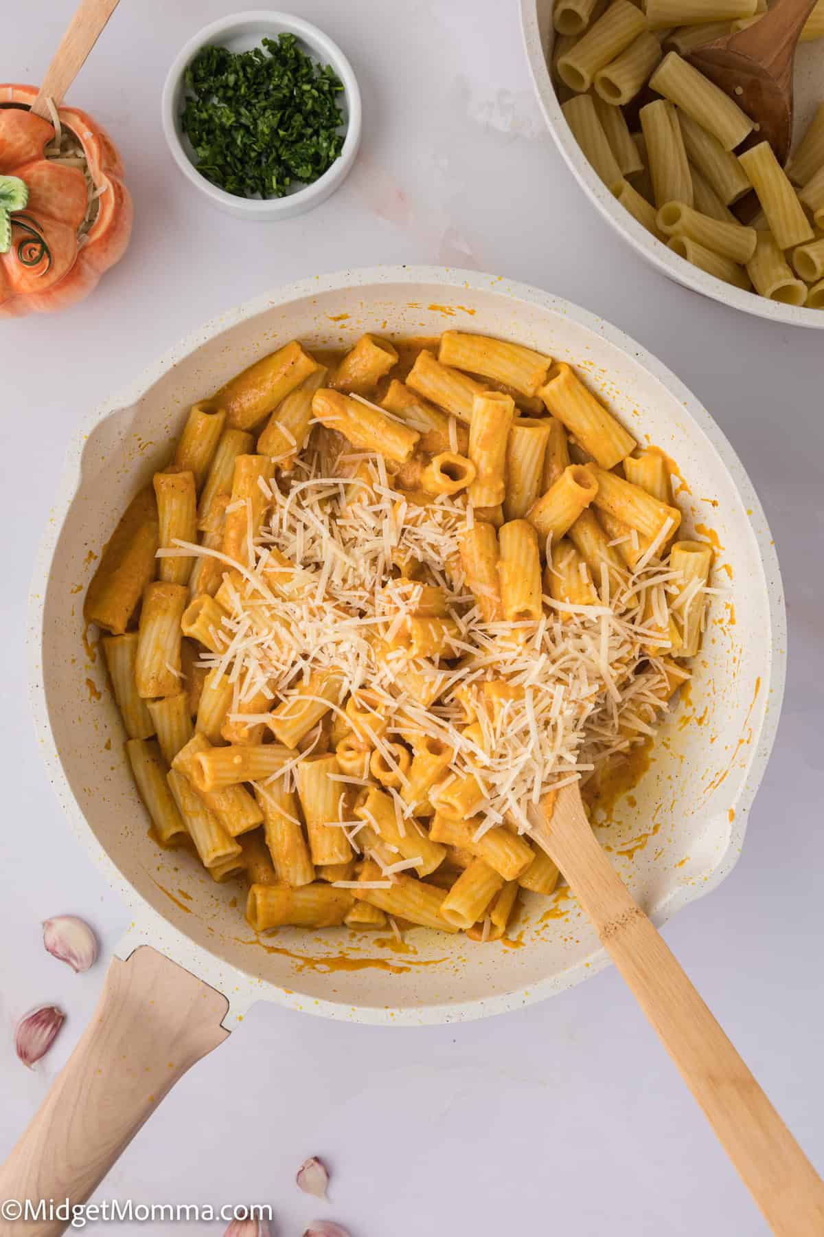 A pot with creamy pumpkin pasta sauce topped with grated cheese, next to a wooden spoon, surrounded by ingredients including garlic and parsley.