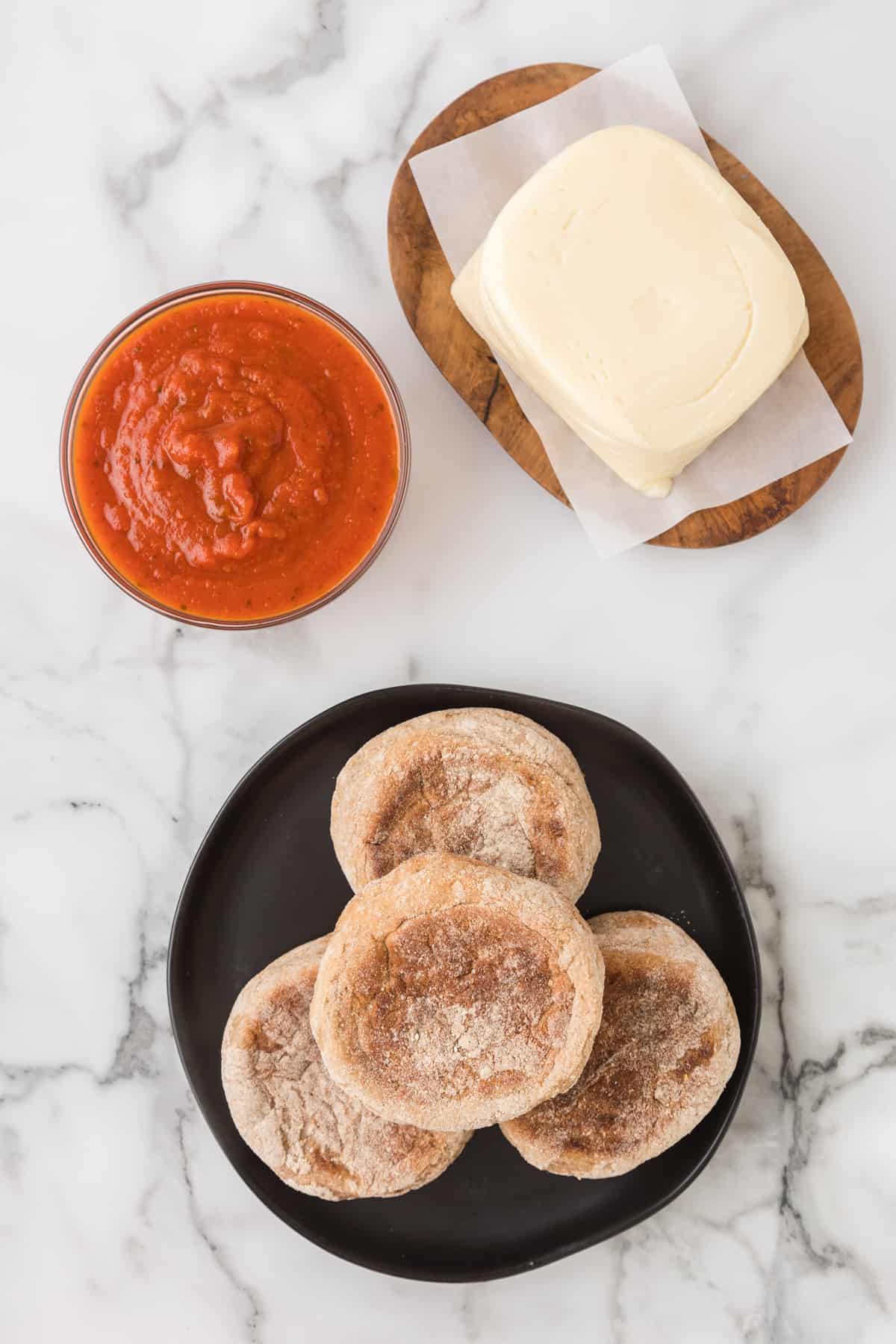 A plate with english muffin, mozzarella cheese and pizza sauce on a marble table.