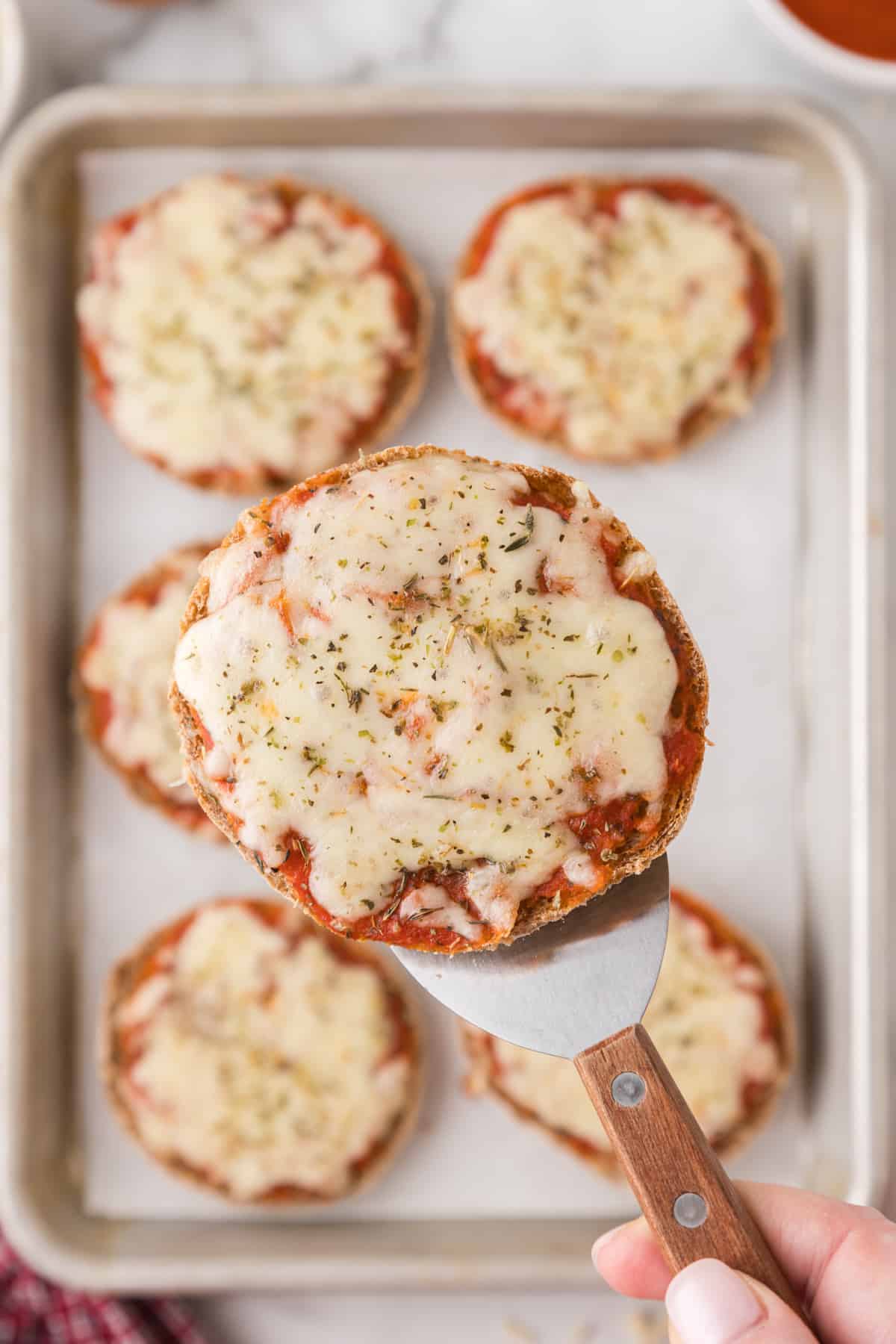 A person is holding a spatula with an english muffin pizza on it. 