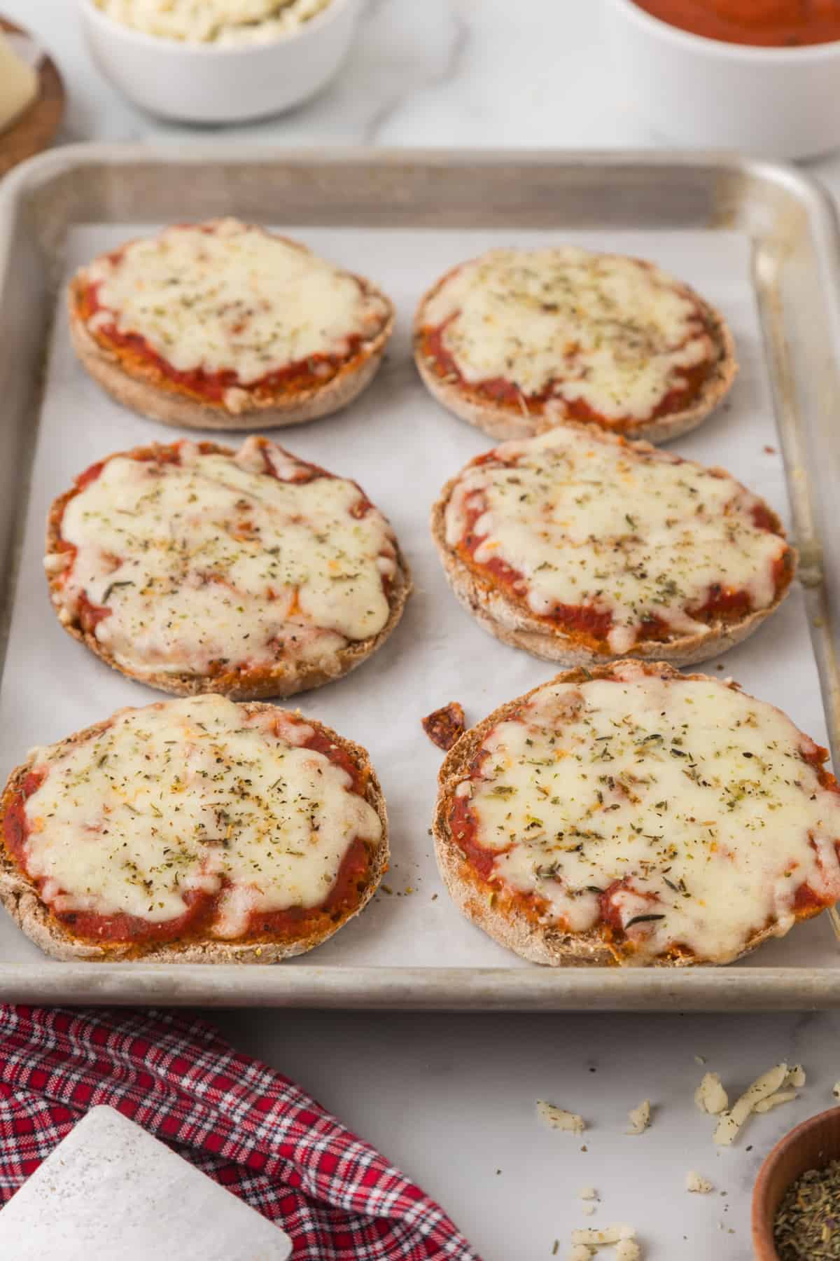 English muffin pizzas with cheese on a baking sheet.