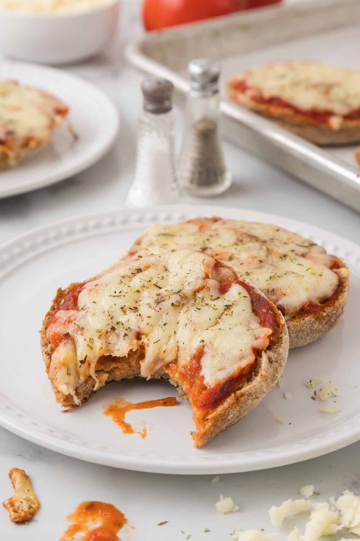 Two halves of English muffin pizza on a plate next to a bowl of sauce.