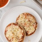 Two halves of English muffin pizza on a plate next to a bowl of sauce.