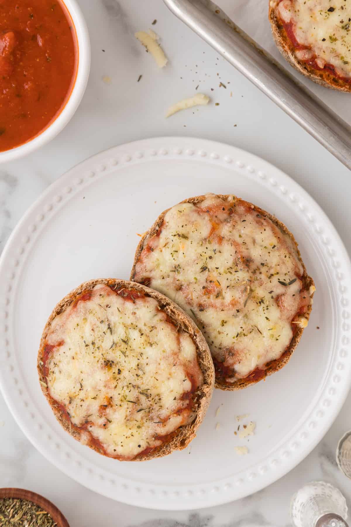 Two halves of English muffin pizza on a plate next to a bowl of sauce.