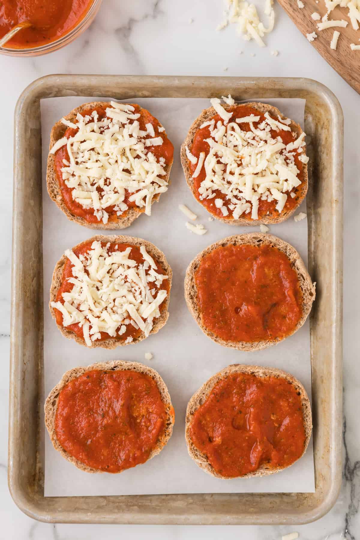 english muffin halves with pizza sauce and shredded mozzarella cheese on a baking sheet.