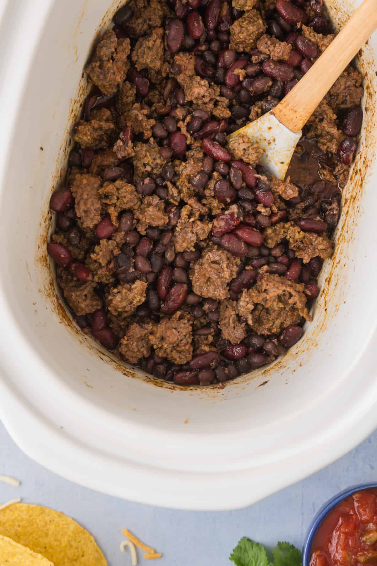 A crock pot full of meat and beans with a wooden spoon.