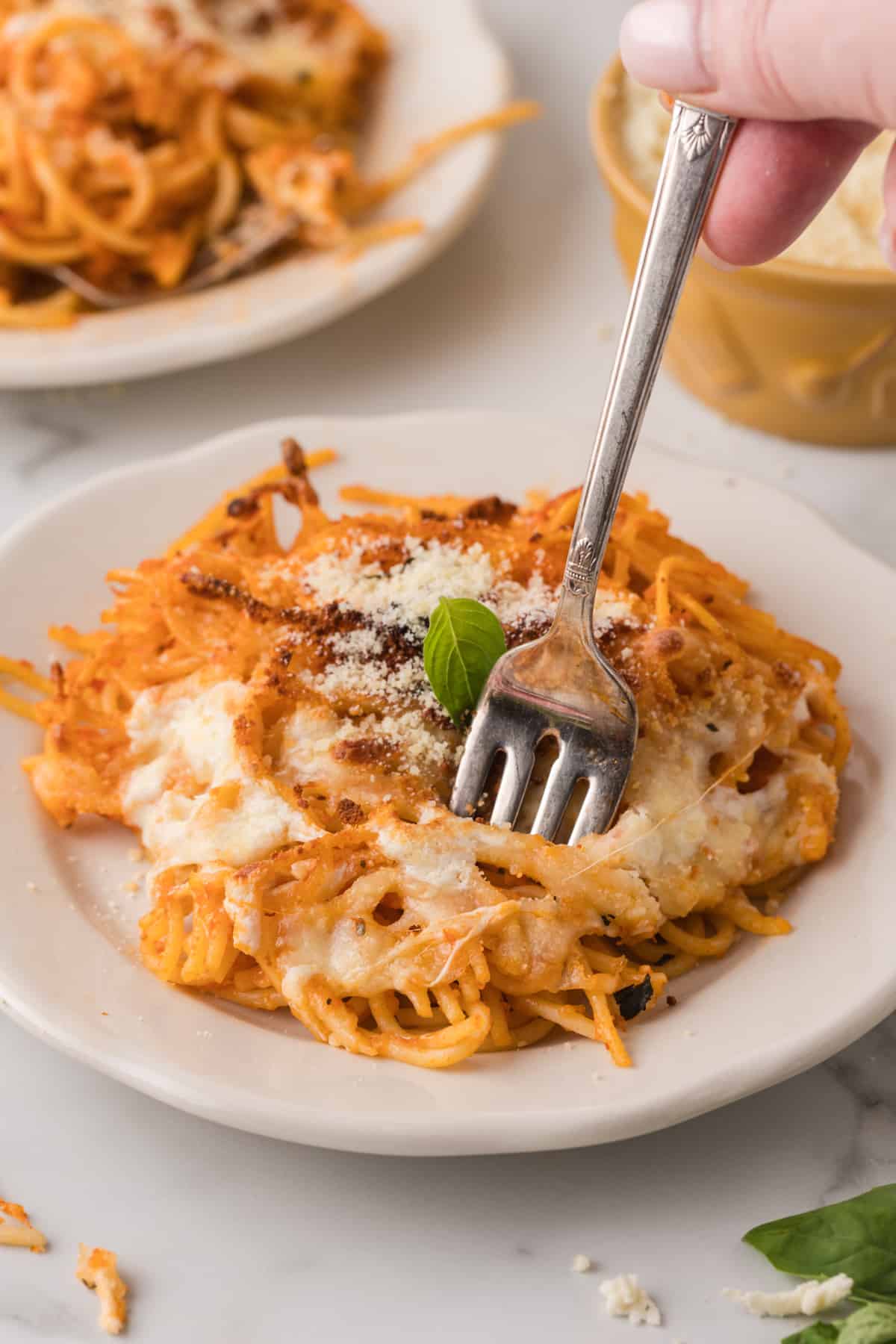 A person holding a fork to a plate of baked spaghetti with melted mozzarella and parmesan cheese.