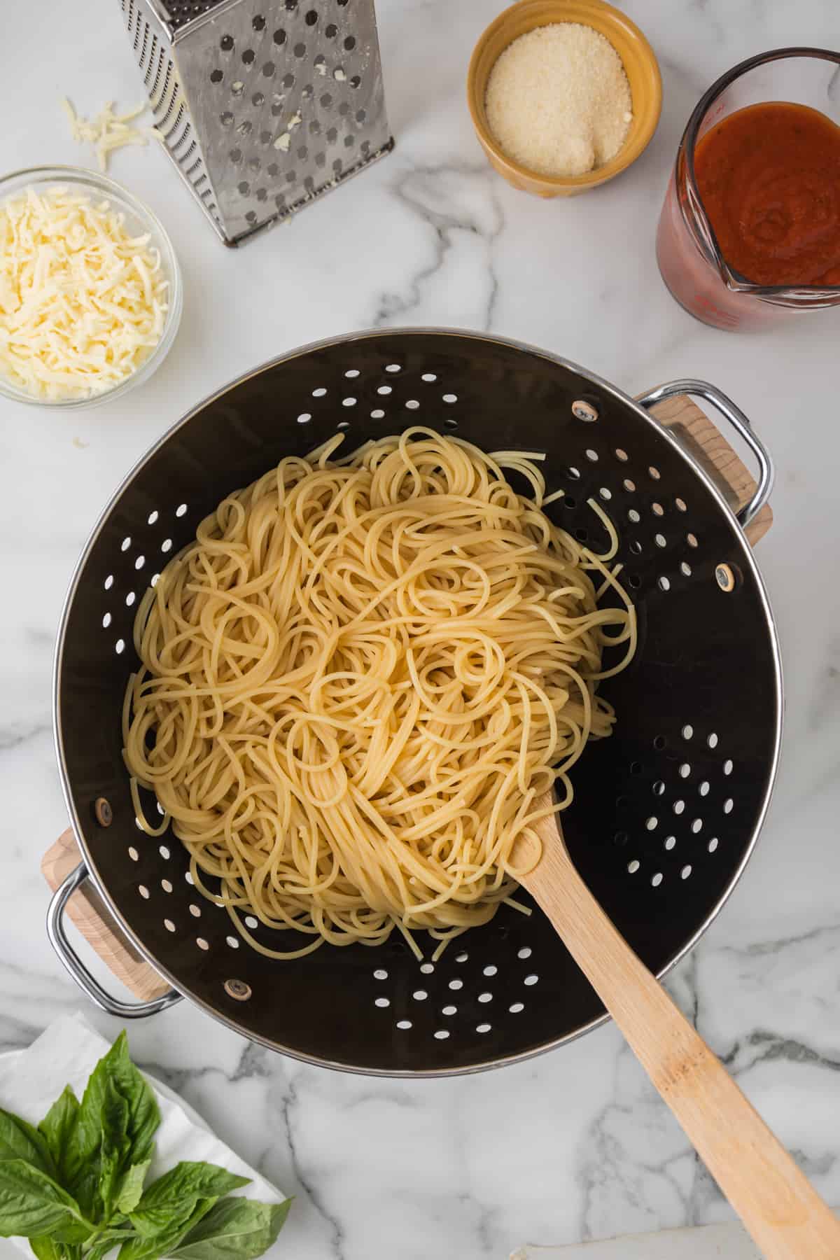 Spaghetti in a colander with ingredients and a wooden spoon.