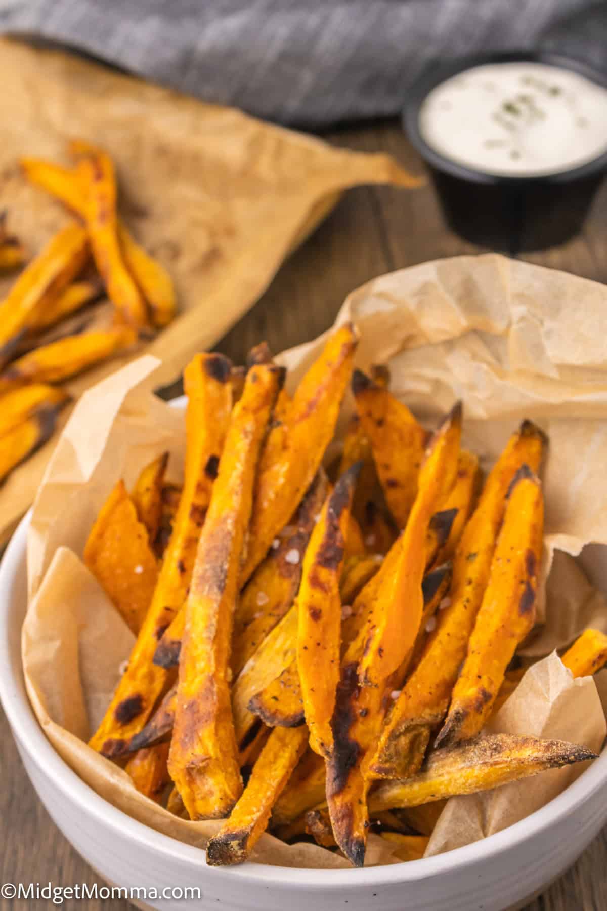 A bowl of baked sweet potato fries on parchment paper with a small dish of dip in the background.