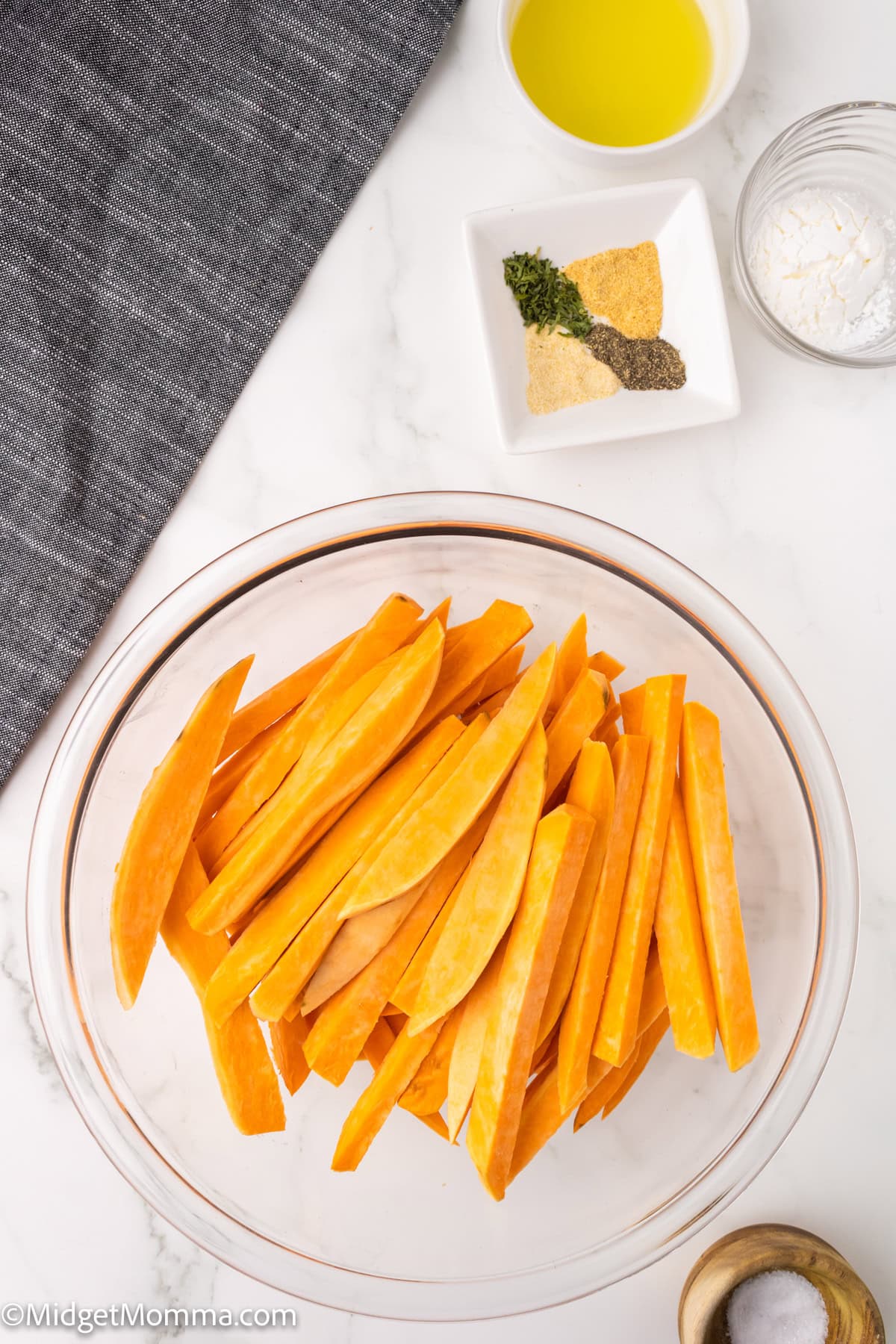 Sweet potato wedges in a glass bowl on a white countertop. Nearby are a small dish with oil, a dish with spices, and a container with cornstarch. A gray cloth is partially visible.