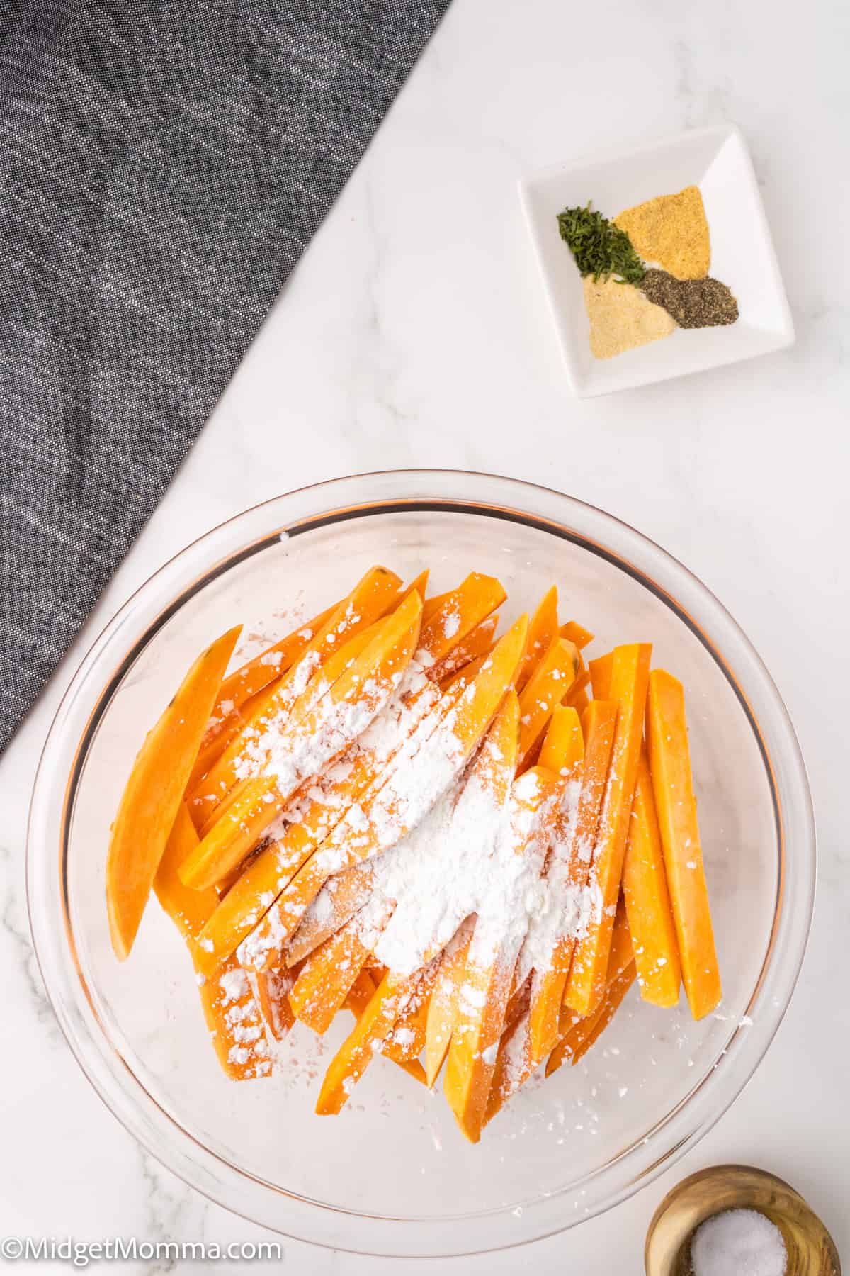 Bowl of raw sweet potato sticks with white powder on top, on a marble surface. Small dish with spices and a wooden object nearby. Dark cloth in the top corner.