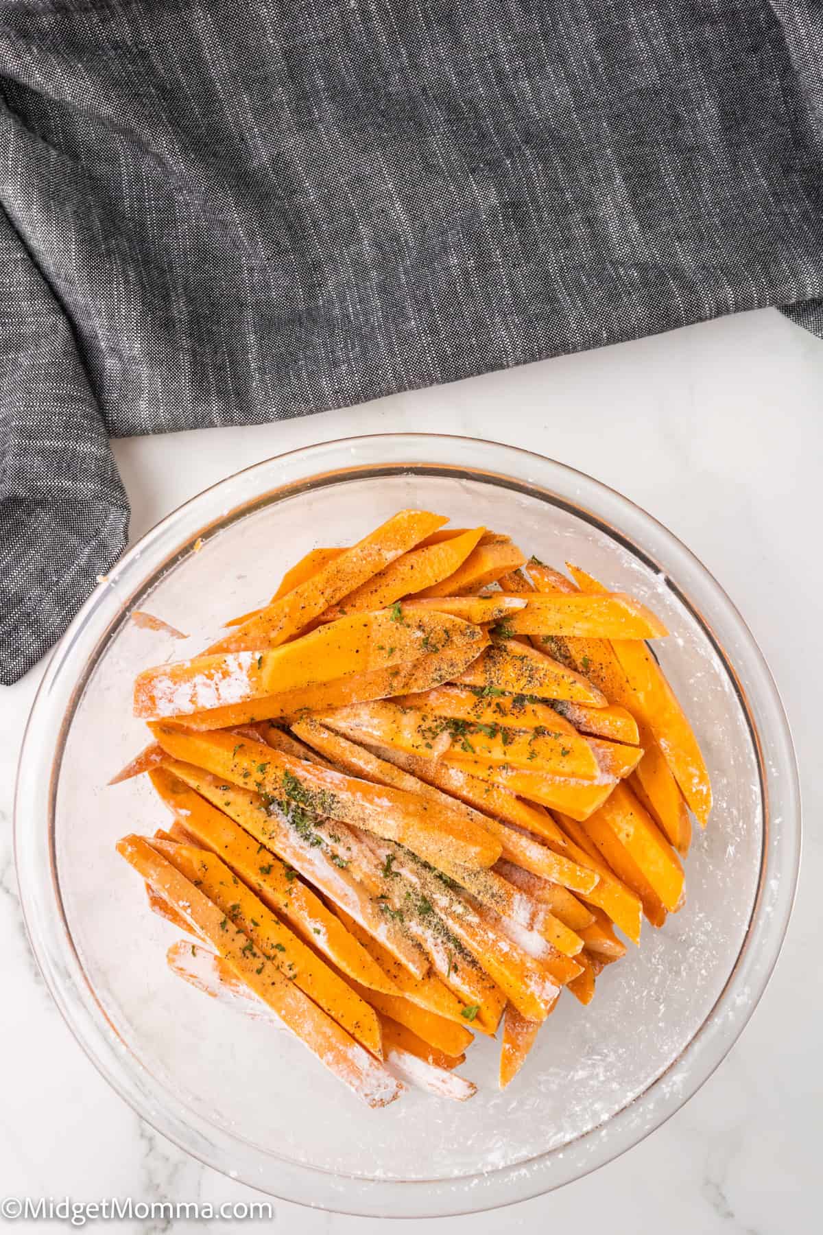 Bowl of uncooked sweet potato fries seasoned with herbs, placed on a countertop with a gray cloth beside it.