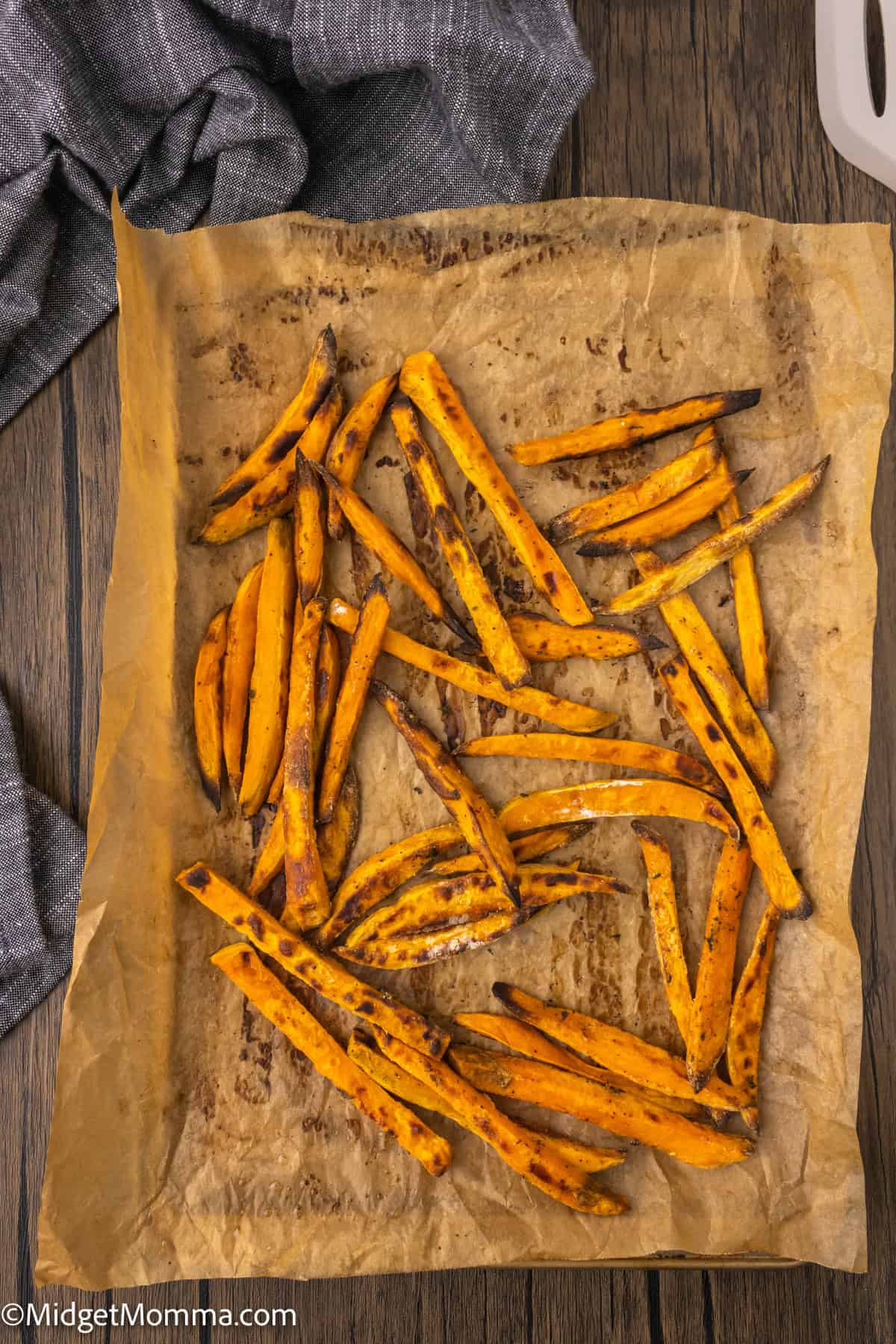 Sweet potato fries on a parchment-lined baking sheet, cooked and slightly charred, with a grey cloth nearby on a wooden surface.