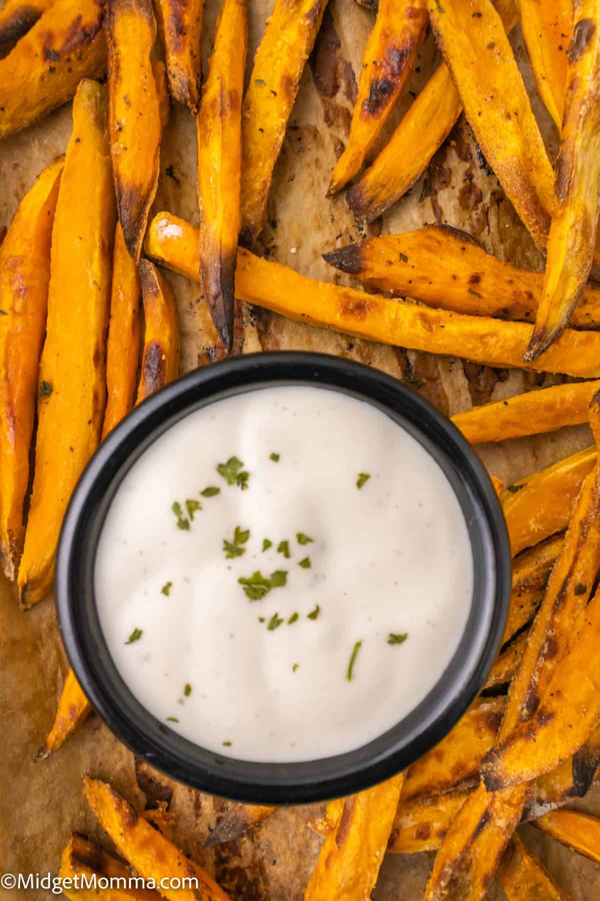 A black bowl of creamy white dipping sauce garnished with herbs, surrounded by baked sweet potato fries on a baking sheet.